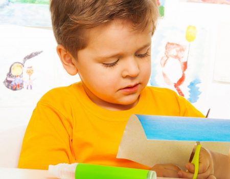 Cute small boy crafting with scissors, paper and glue sitting at the table with drawings on background